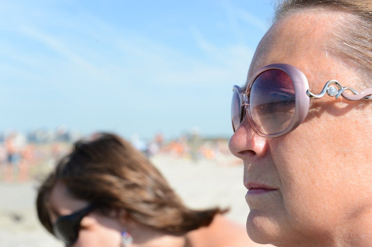 A woman wearing large sunglasses looks into the distance at the beach, with another person slightly out of focus.