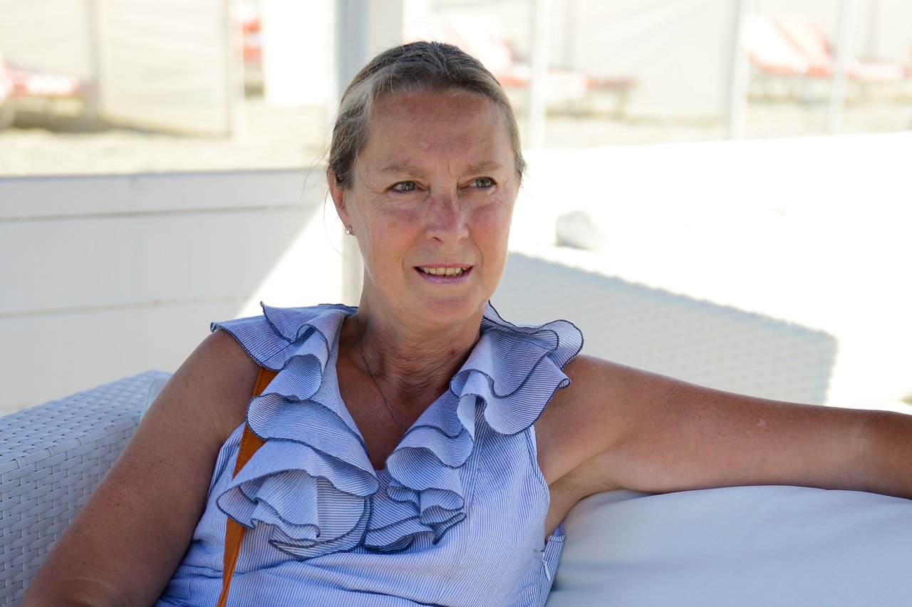 A woman sits on a white outdoor couch, wearing a blue ruffled top and looking slightly to the side.