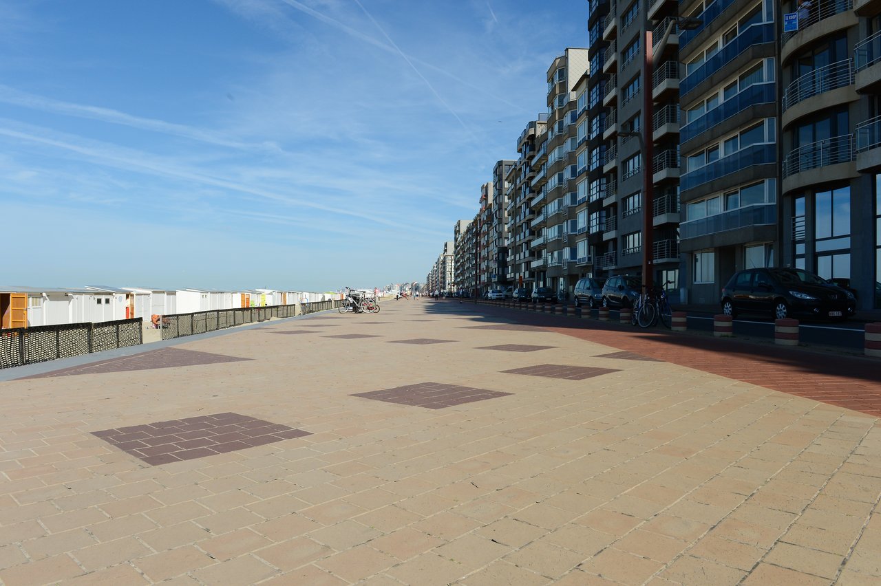 A wide promenade with beachfront cabins on one side and tall apartment buildings with parked cars on the other.