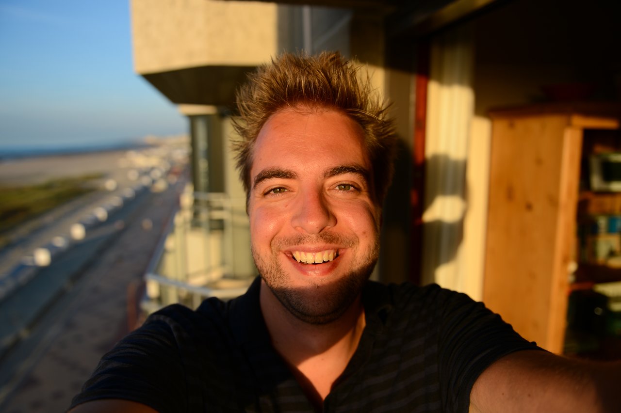 A person takes a smiling selfie on a balcony with sunlight on their face and a beach in the background.