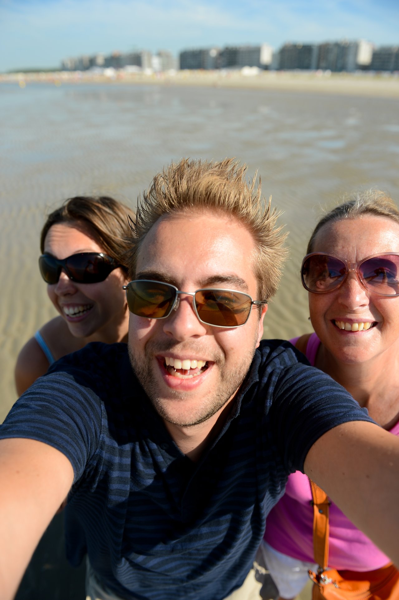 A man takes a selfie with two smiling women on a sandy beach with buildings in the background.