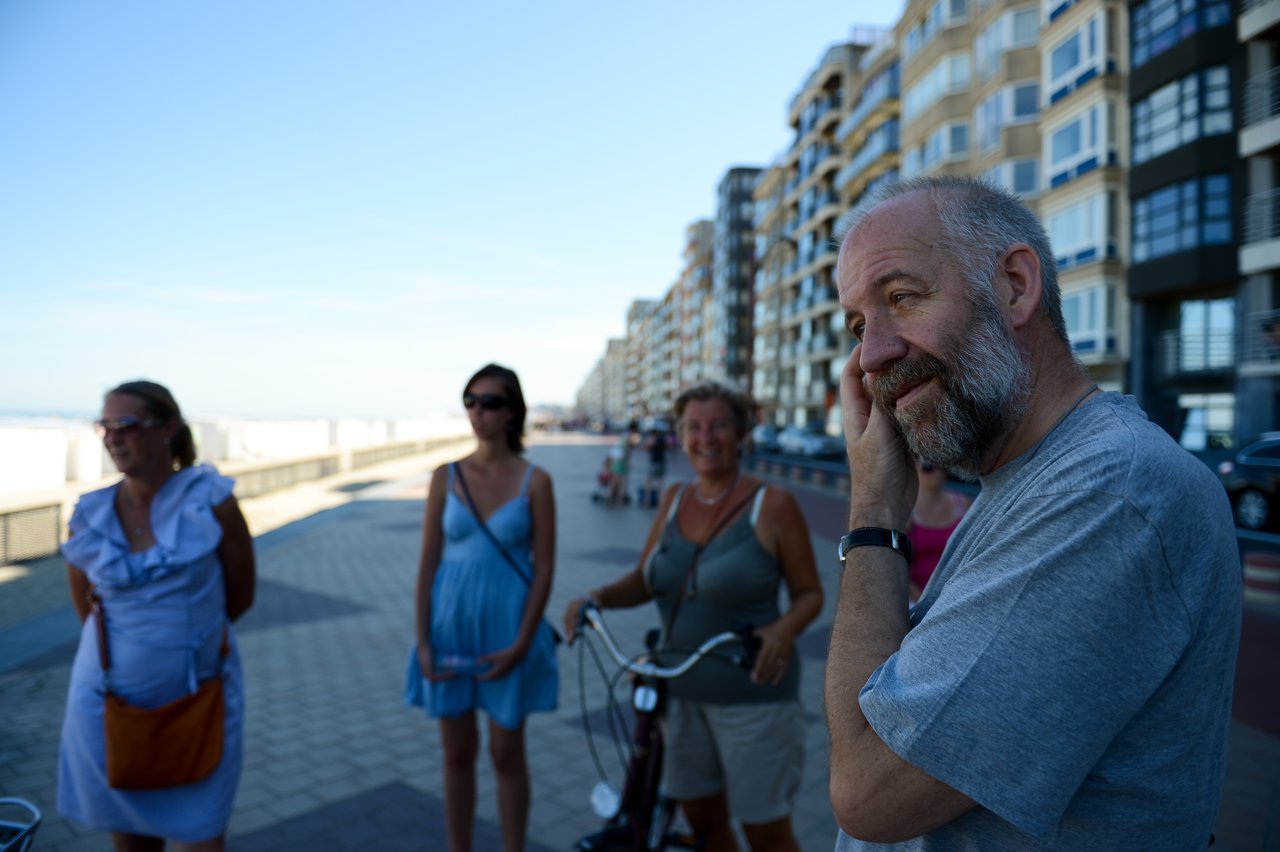 A bearded man in a gray shirt talks while touching his face, with three women listening nearby.