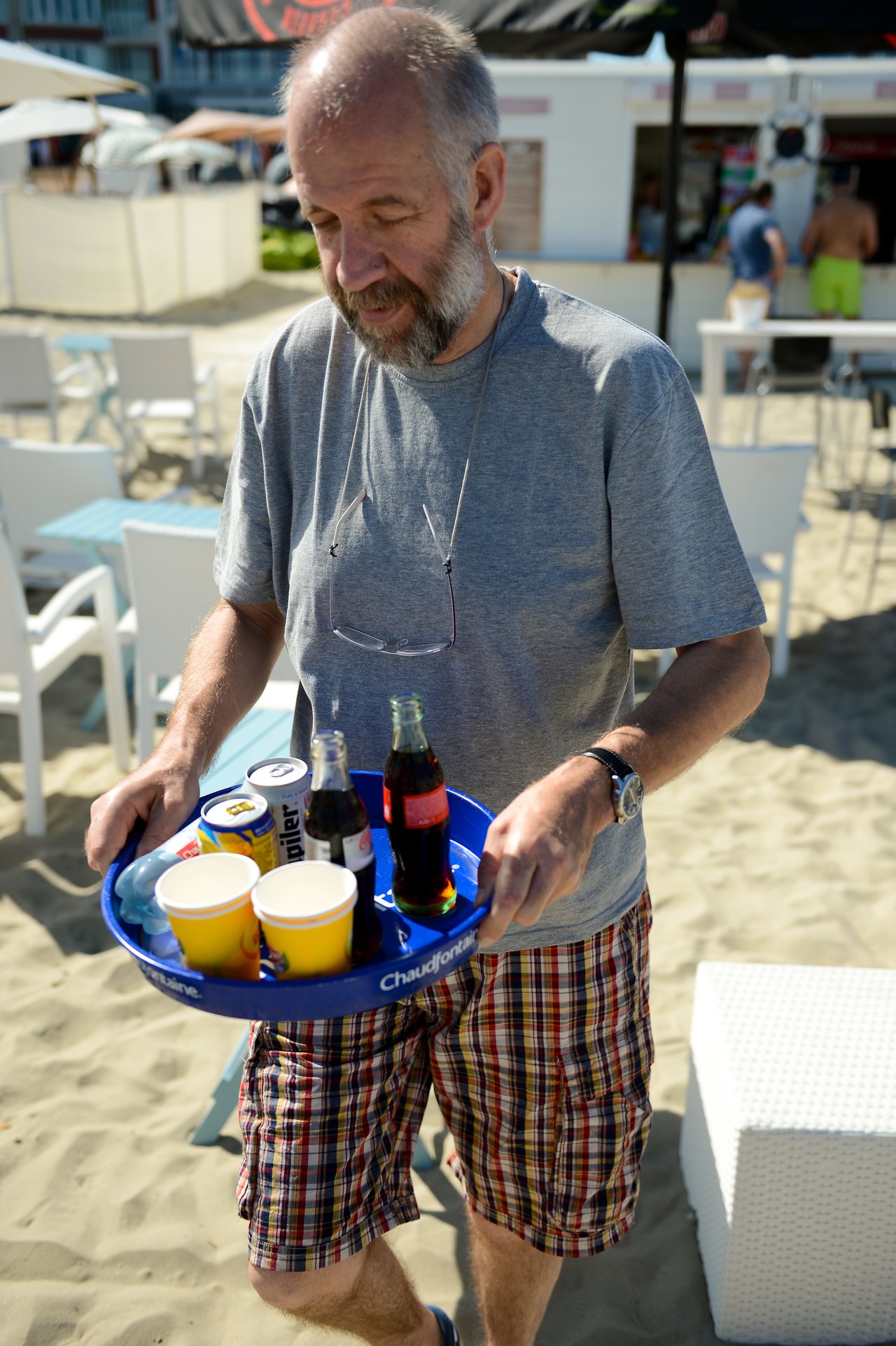 A man in casual clothes carries a tray with drinks on a sandy beach, walking towards a seating area.