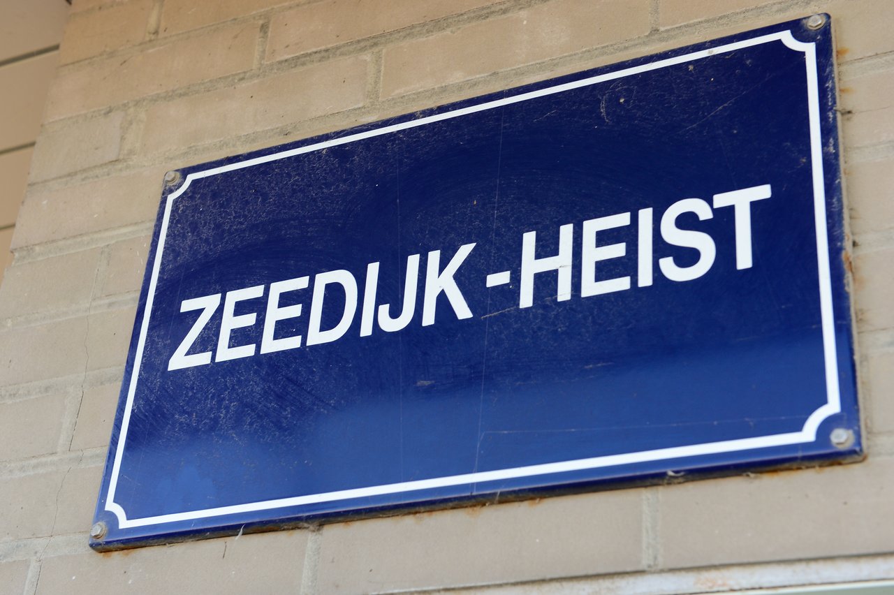 A blue street sign with white text reads "ZEEDIJK-HEIST", mounted on a brick wall.