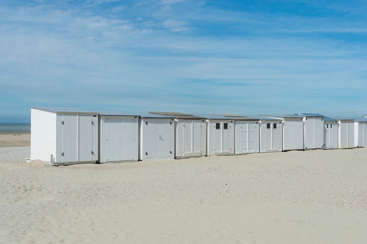 A row of small white beach huts stands on a sandy shore under a clear blue sky.