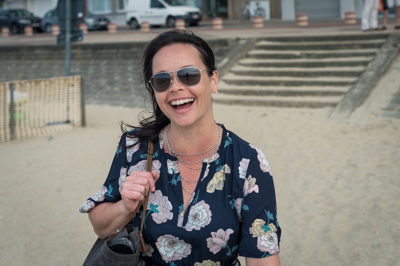A smiling woman wearing sunglasses and a floral dress walks on a sandy beach, carrying a bag on her shoulder.