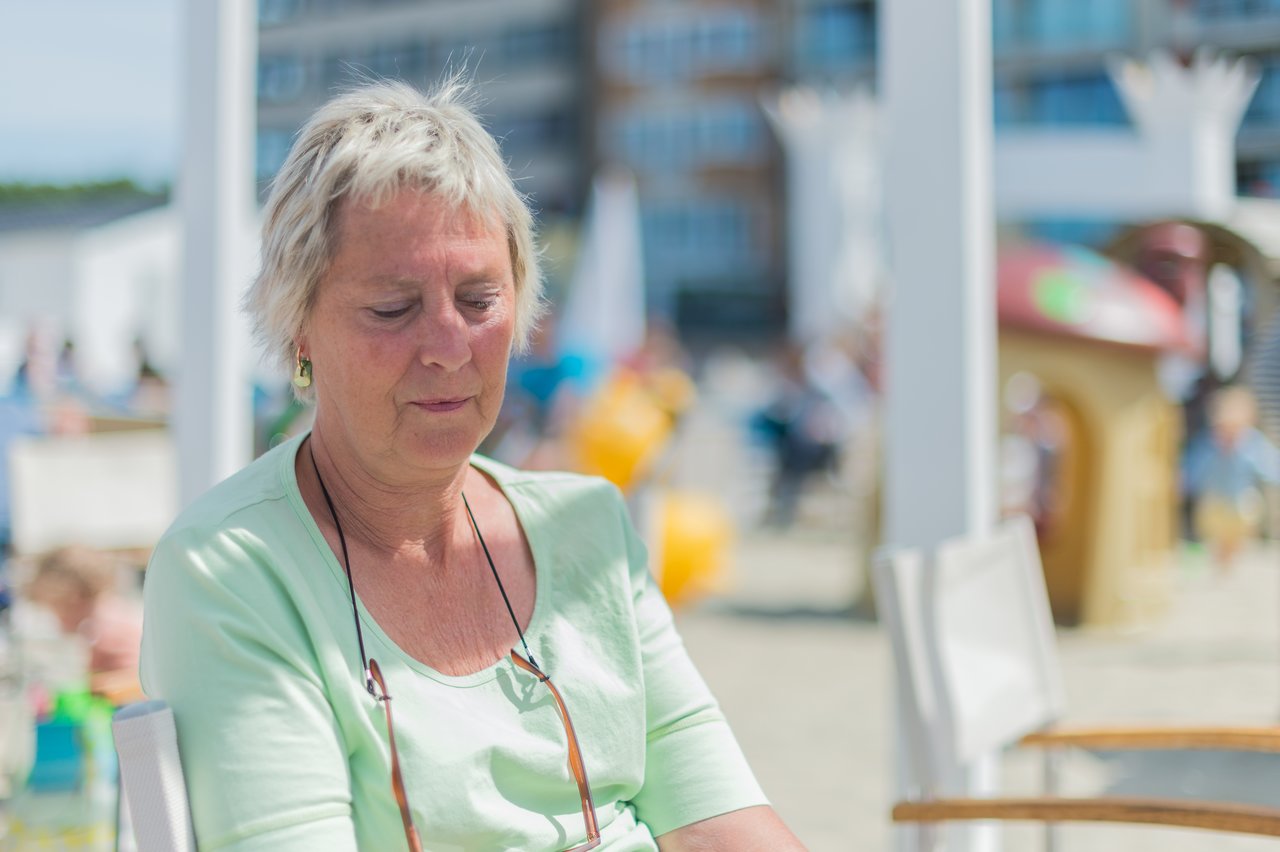 An older woman in a light green shirt sits outdoors with her eyes closed.