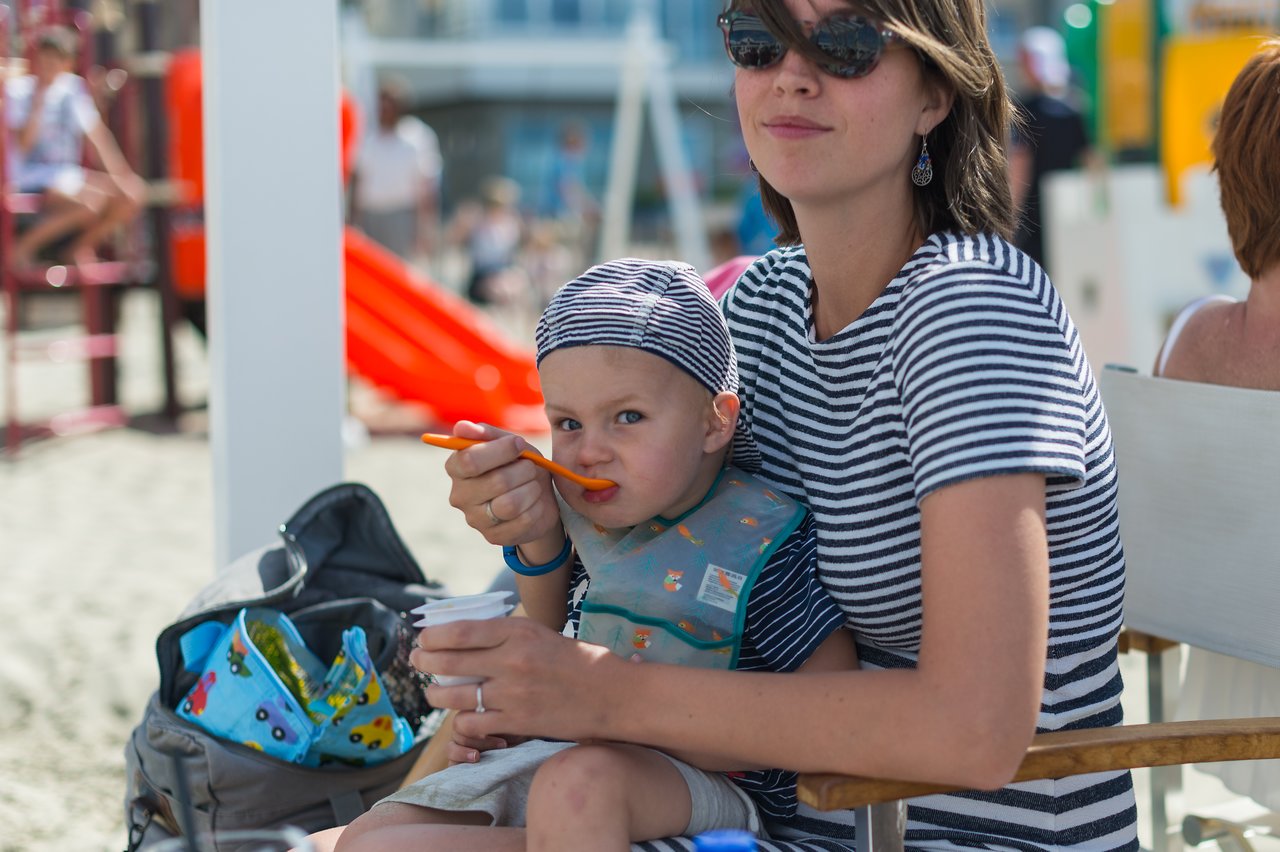 A woman in striped clothing feeds a young child with a spoon while sitting outdoors.