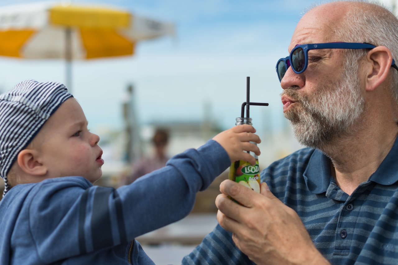 A child holds a juice bottle with a straw while an older man playfully puckers his lips to drink.