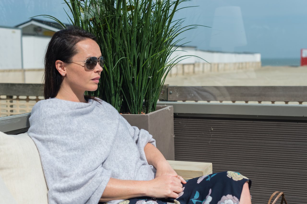 A woman wearing sunglasses sits on an outdoor couch, looking into the distance with a relaxed expression.