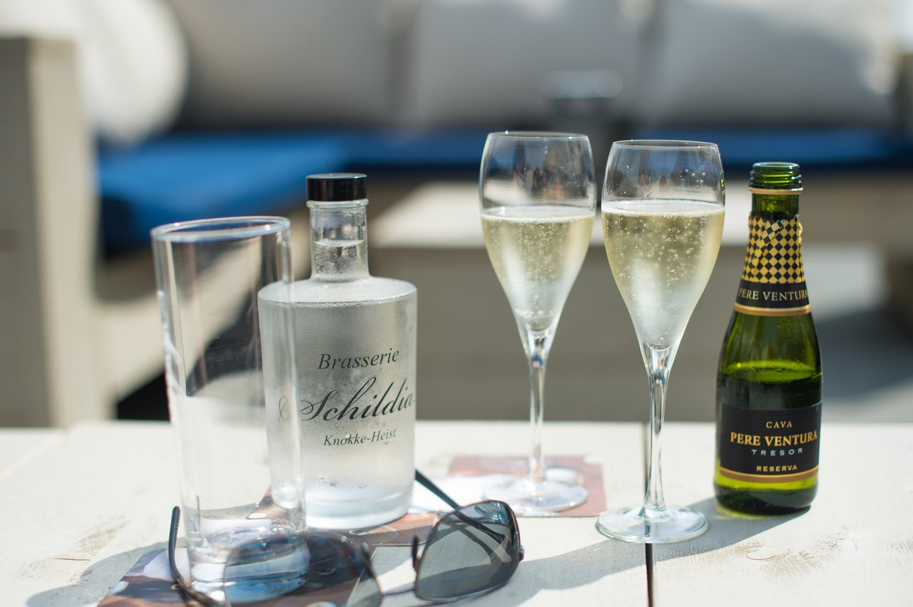 Two glasses of sparkling wine, a bottle of cava, a water bottle, and sunglasses on a table.