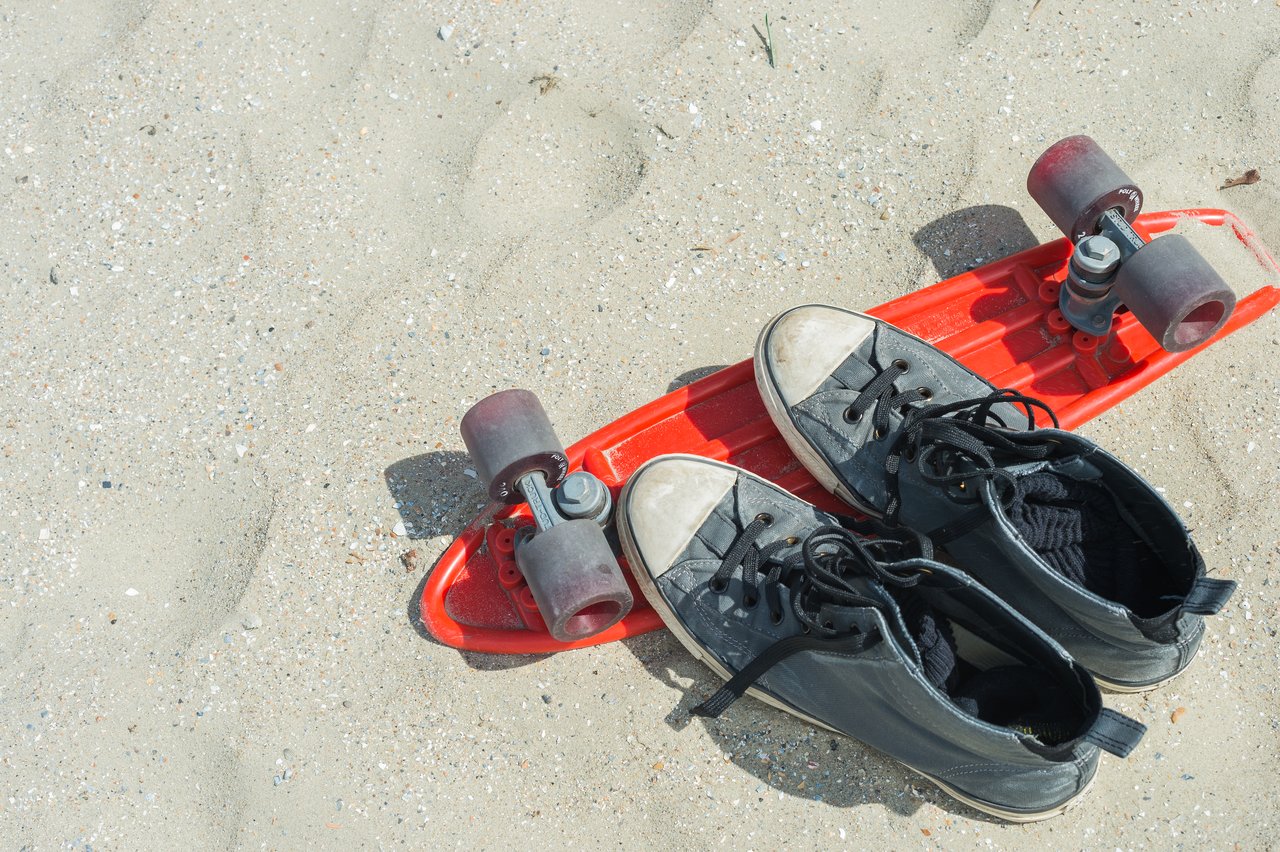 A pair of black sneakers rests on a red skateboard placed on a sandy beach.