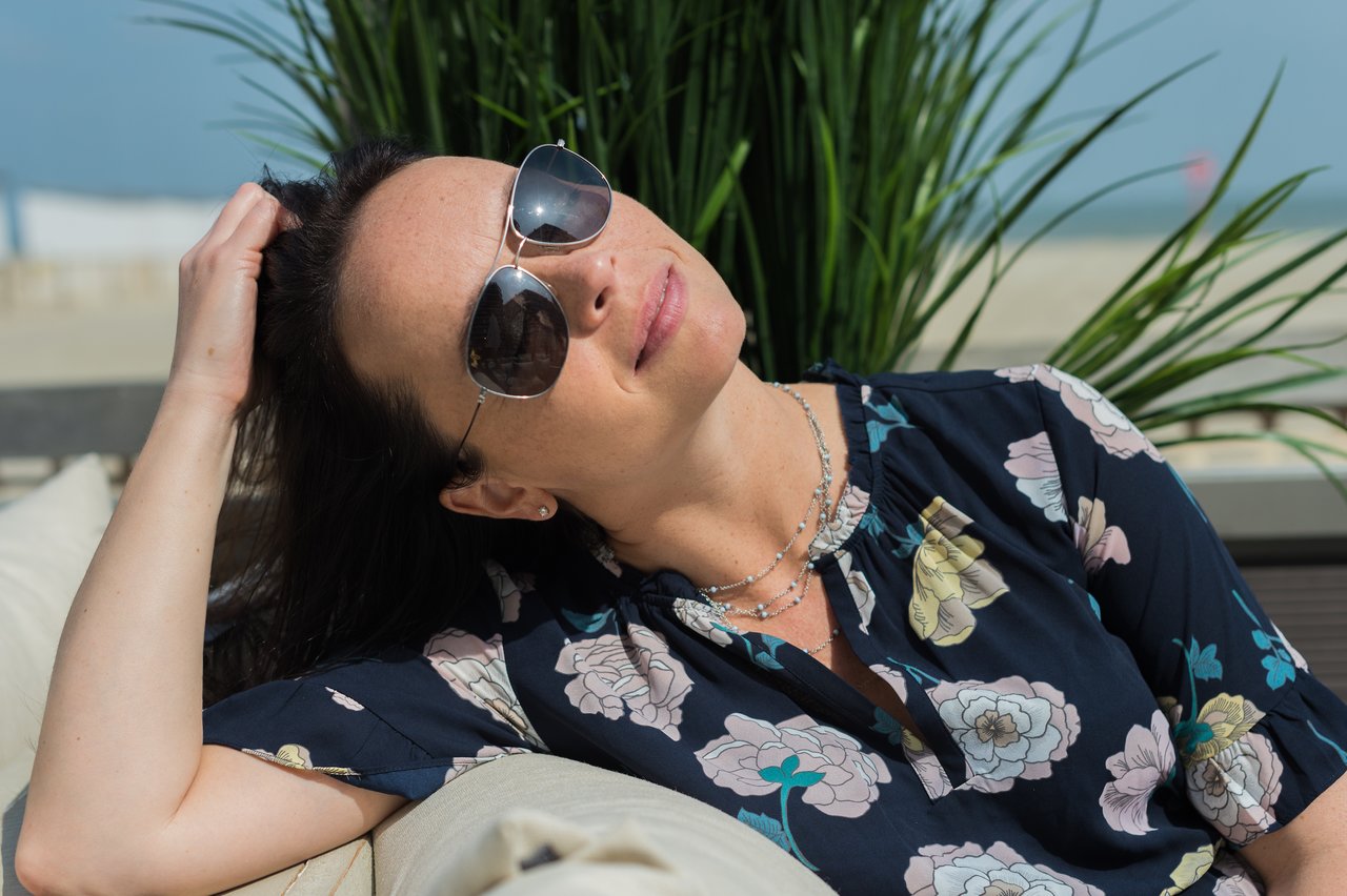 A woman wearing sunglasses and a floral blouse leans back on a couch, resting her head with a relaxed expression.