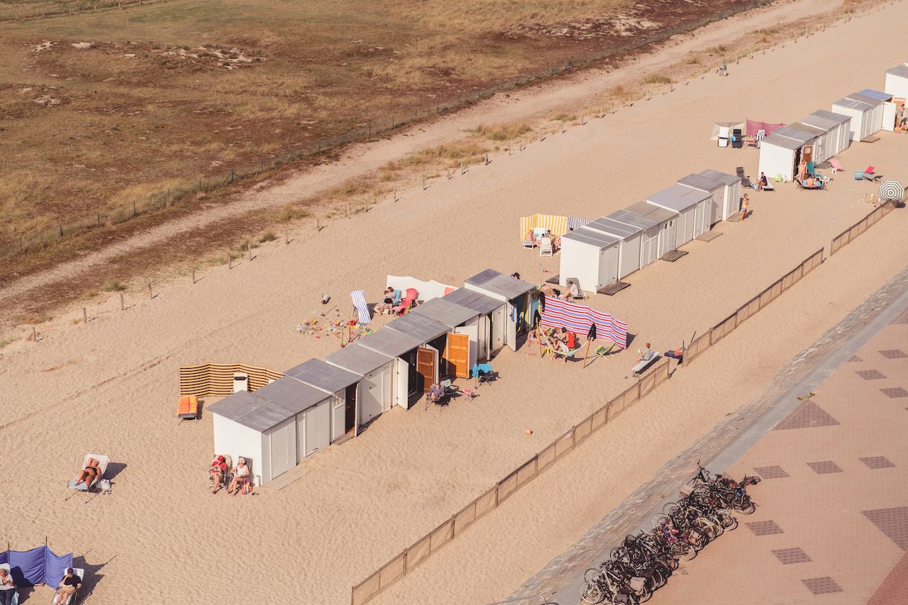 Rows of small beach huts on sandy shore with people relaxing, sunbathing, and sitting under umbrellas.