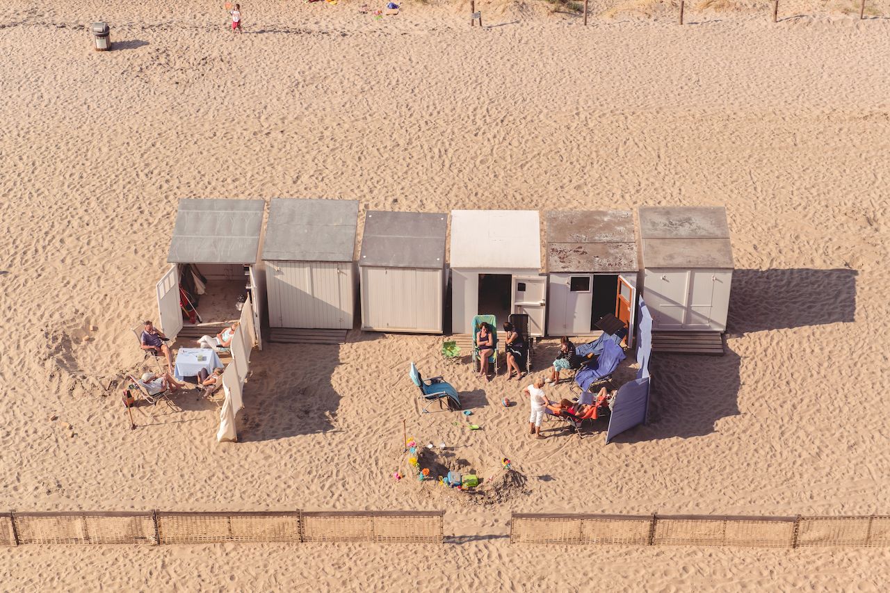 A row of beach huts with people sitting outside on chairs, relaxing and socializing on the sandy beach.