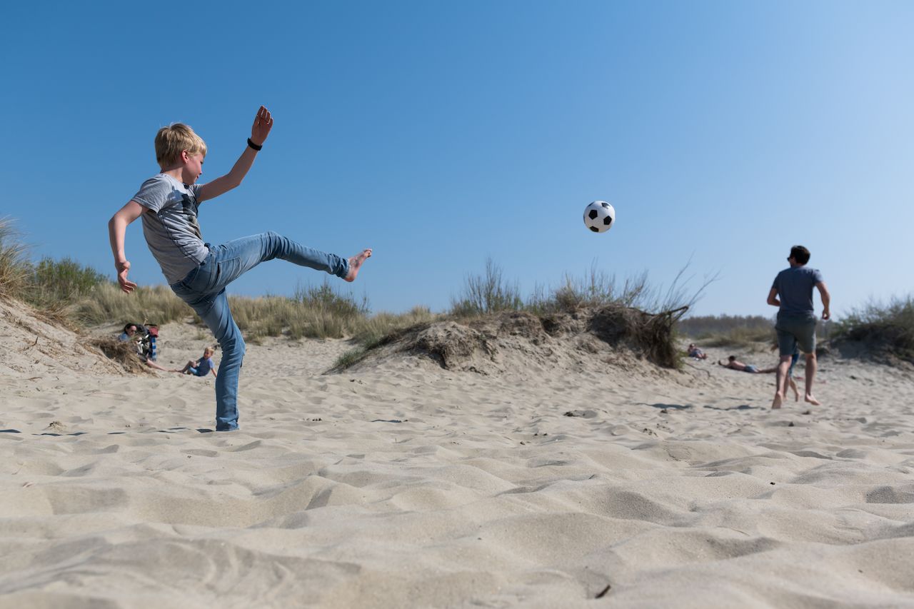 A boy kicks a soccer ball in the sandy dunes while another person runs toward it.