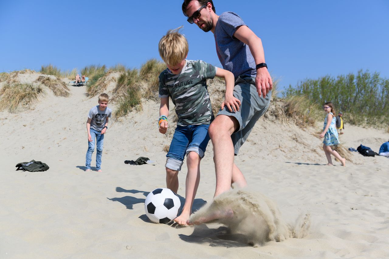 A child and an adult play soccer on sandy dunes, kicking up sand as they compete for the ball.