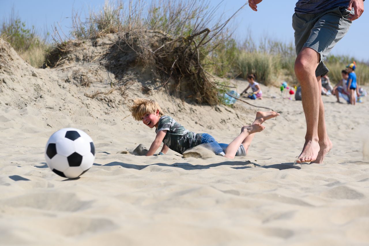 A child falls in the sand while chasing a soccer ball, as an adult runs nearby.