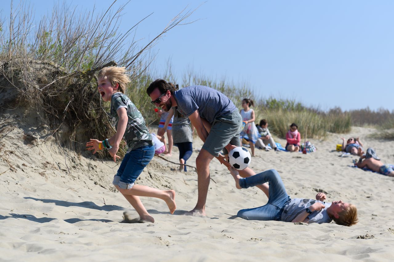 A man and two children play soccer on sandy dunes, with one child falling while kicking the ball.
