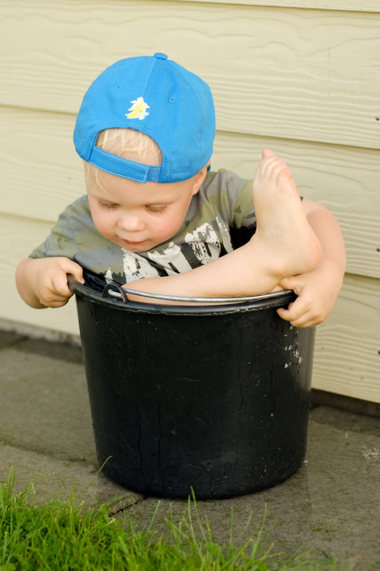 A young child wearing a blue cap sits inside a black bucket, holding the edges and lifting one leg.