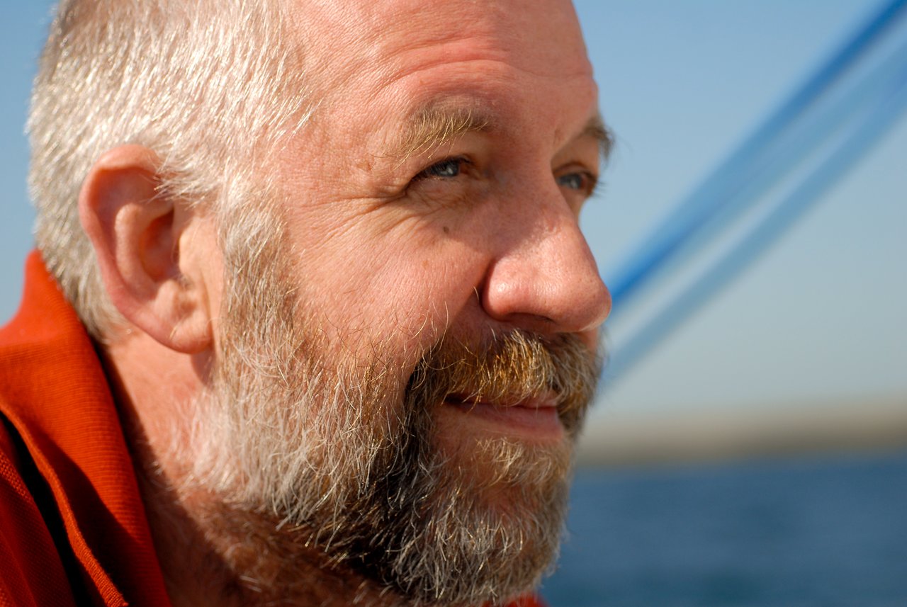 A bearded man in a red shirt smiles while looking into the distance, with a blurred blue sky behind him.