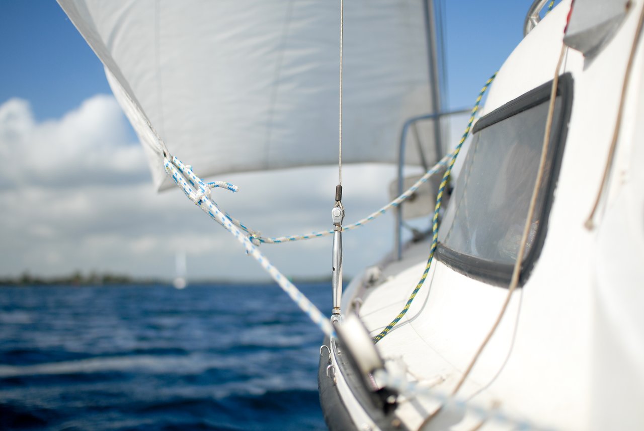 Close-up of a sailboat's rigging and ropes with the sail raised, moving through the water under a partly cloudy sky.