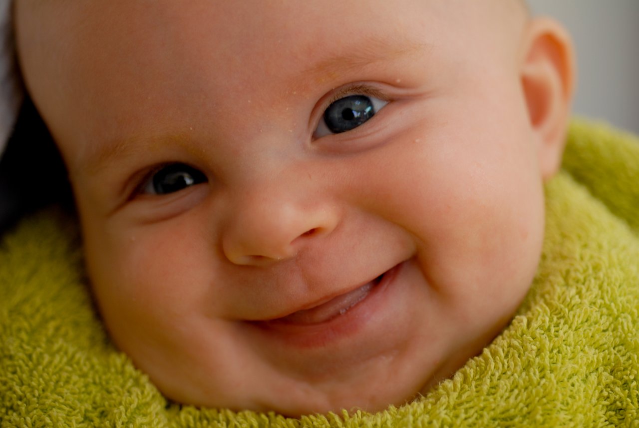 A baby wrapped in a green towel smiles with bright eyes and a slightly open mouth.