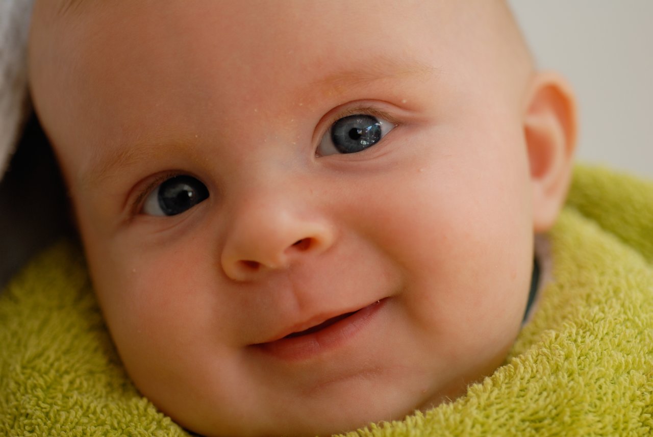 A close-up of a baby wrapped in a green towel, smiling and looking directly at the camera.