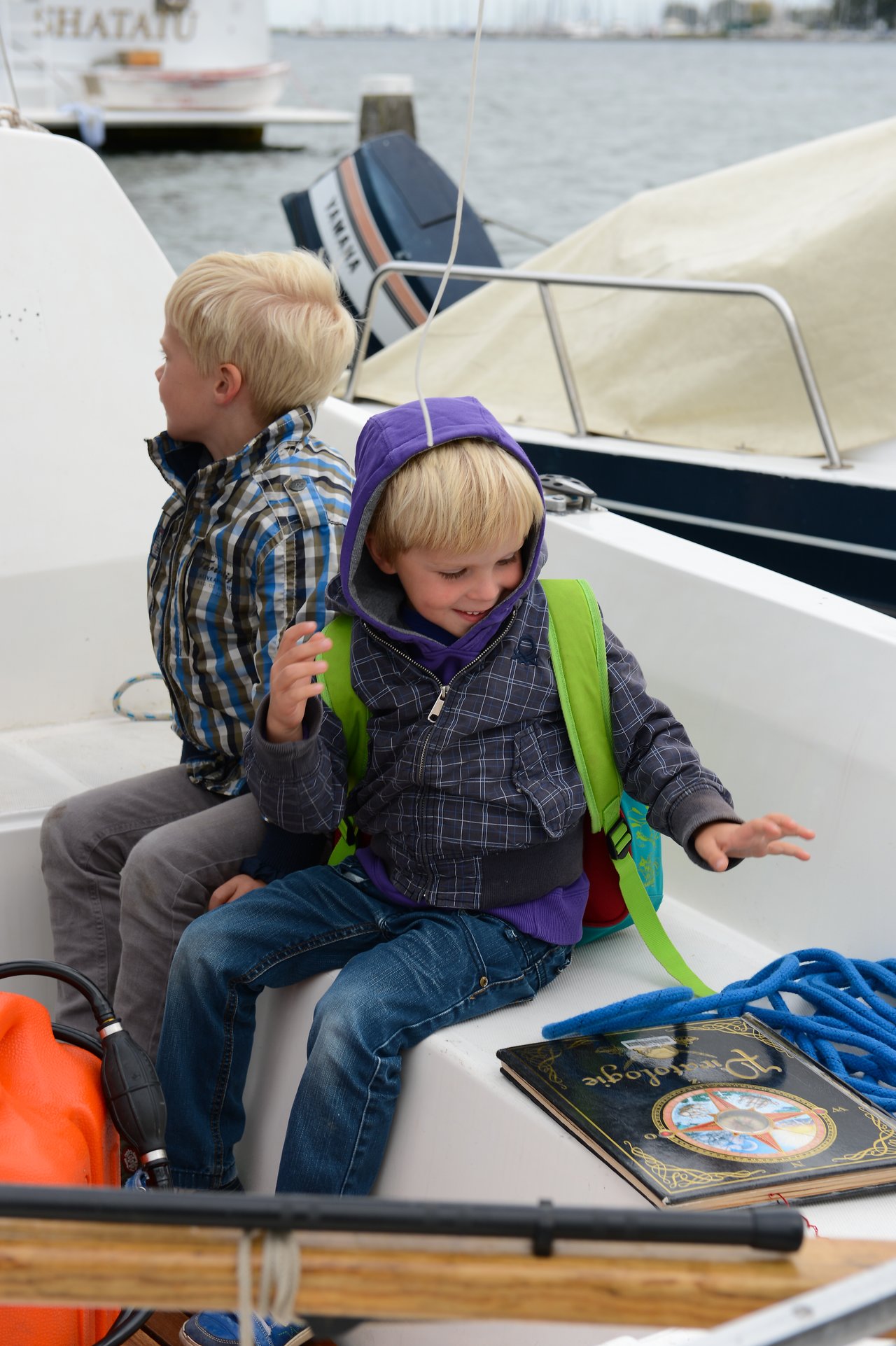 Two young children with backpacks sit on a boat, one smiling and moving excitedly while the other looks away.