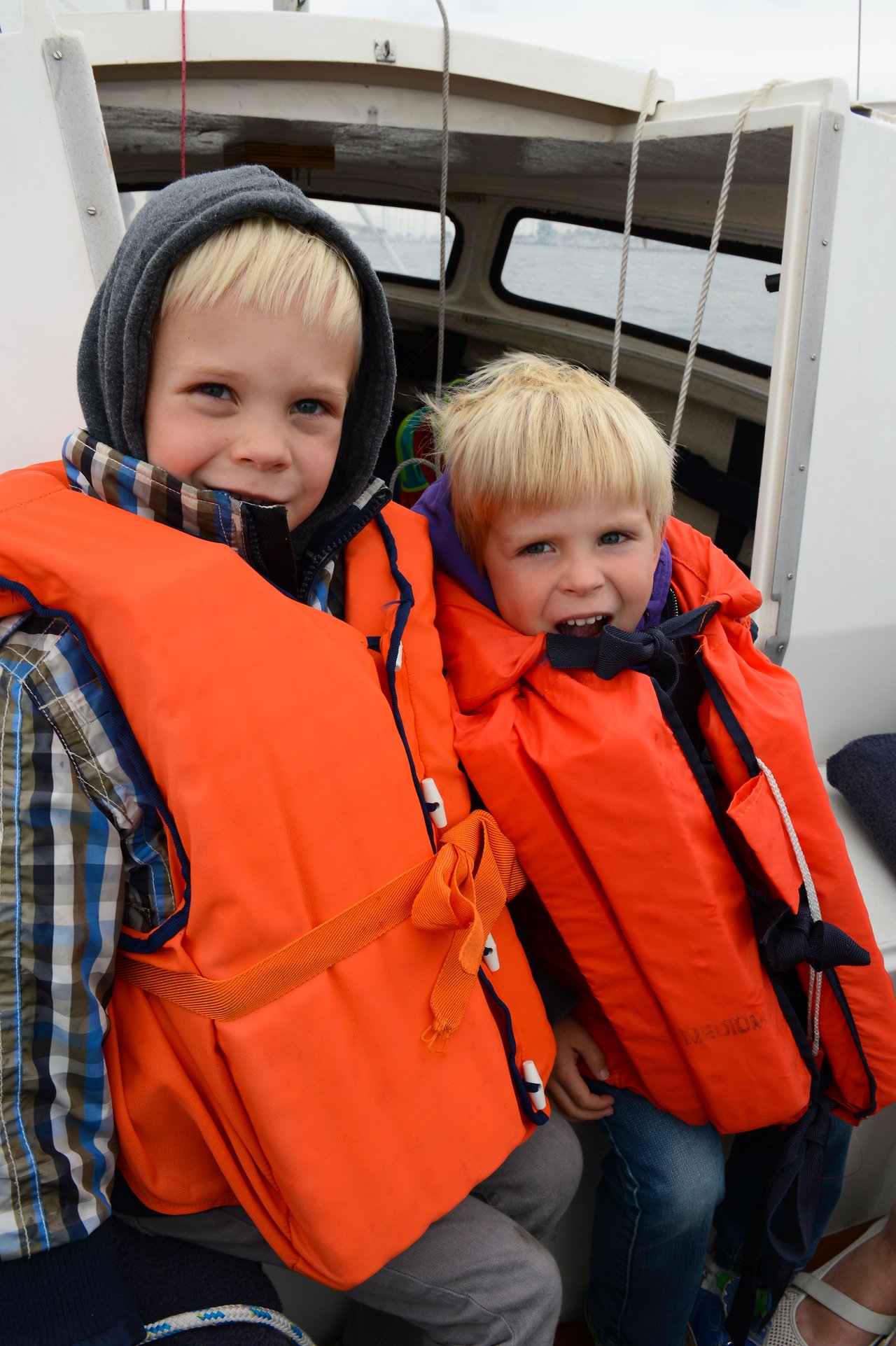 Two young children wearing bright orange life jackets sit on a boat, smiling at the camera.