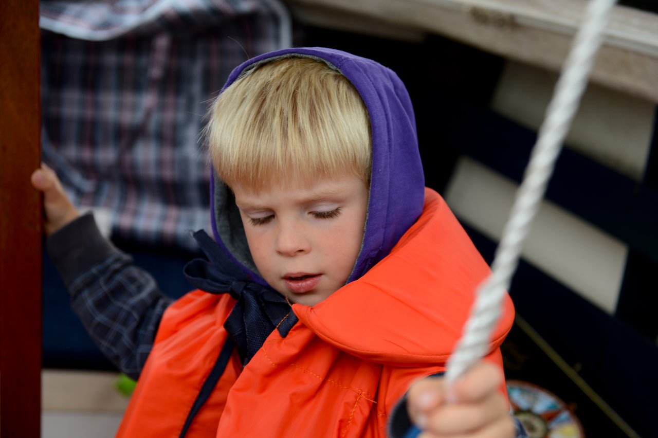 A young child wearing a life jacket and hoodie holds onto a rope while looking down.