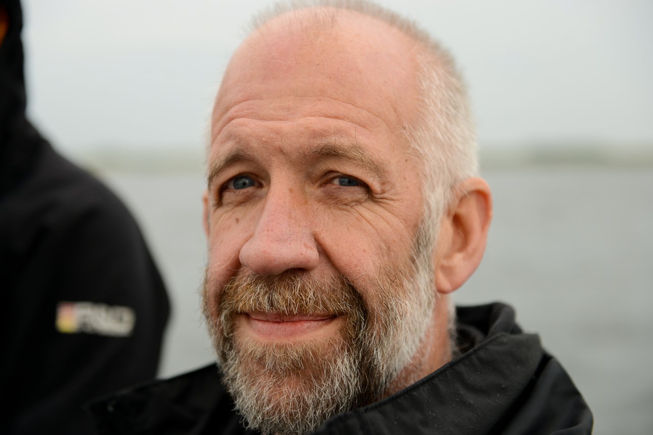 A bearded man in a black jacket smiles slightly while looking at the camera, with water in the background.