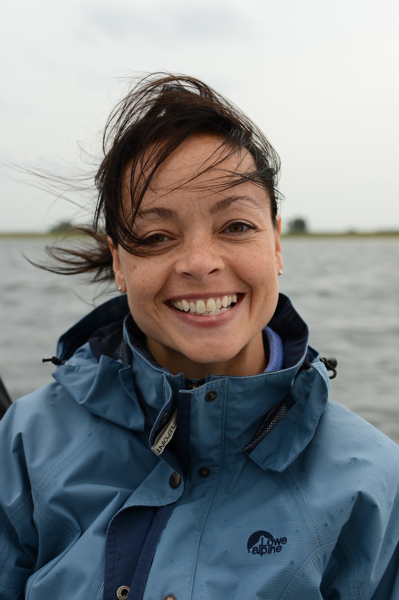 A smiling person in a blue jacket outdoors with windblown hair, standing near a body of water.