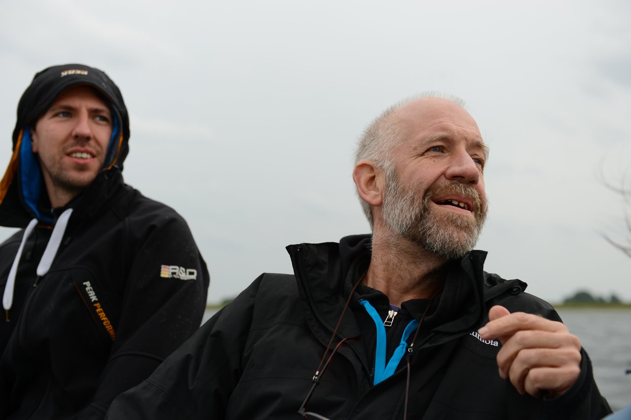 Two men in jackets sit outdoors, one smiling and gesturing while the other looks in the same direction.