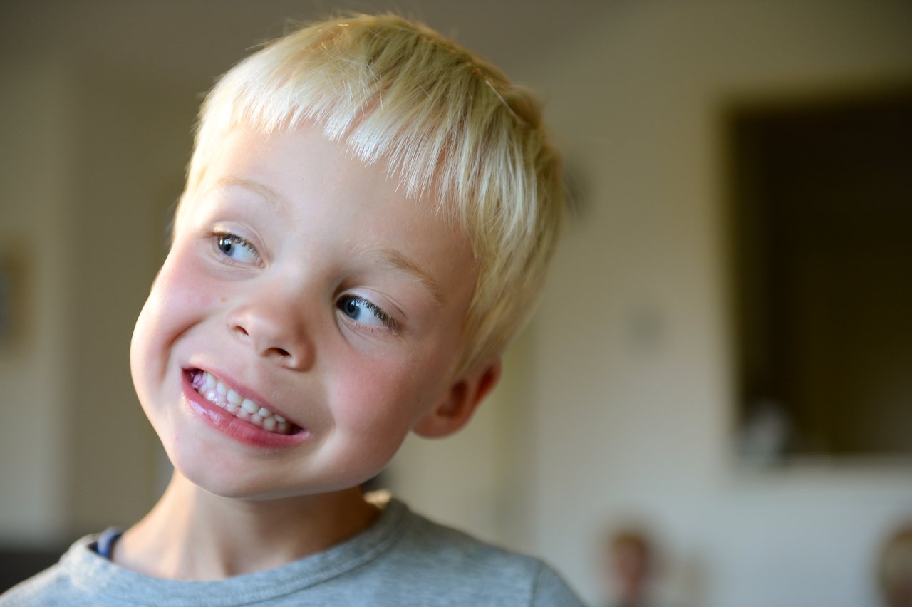 A young child with blonde hair tilts their head and smiles, showing their teeth.