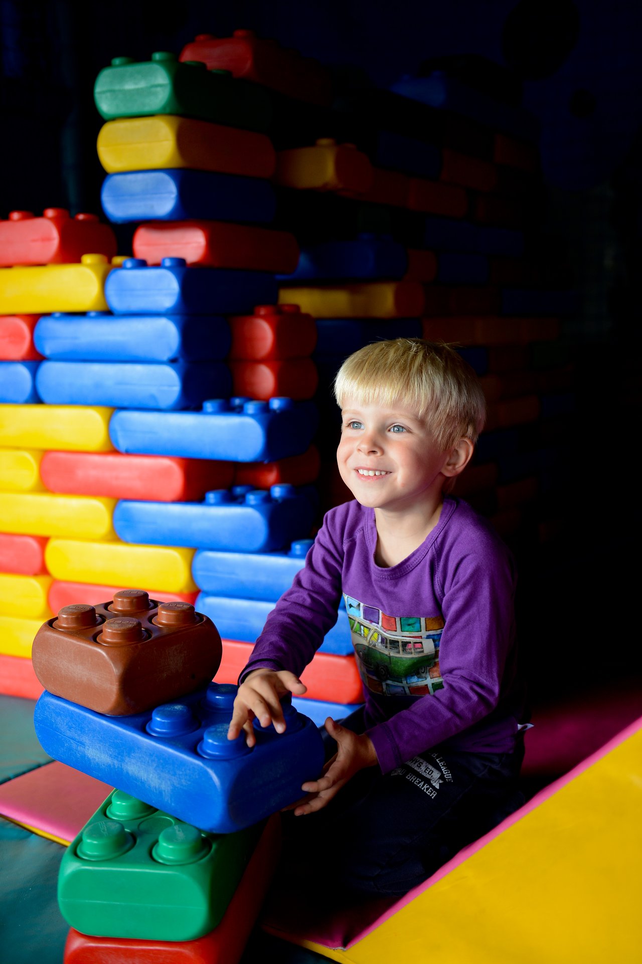 A smiling child in a purple shirt plays with large, colorful building blocks in an indoor play area.