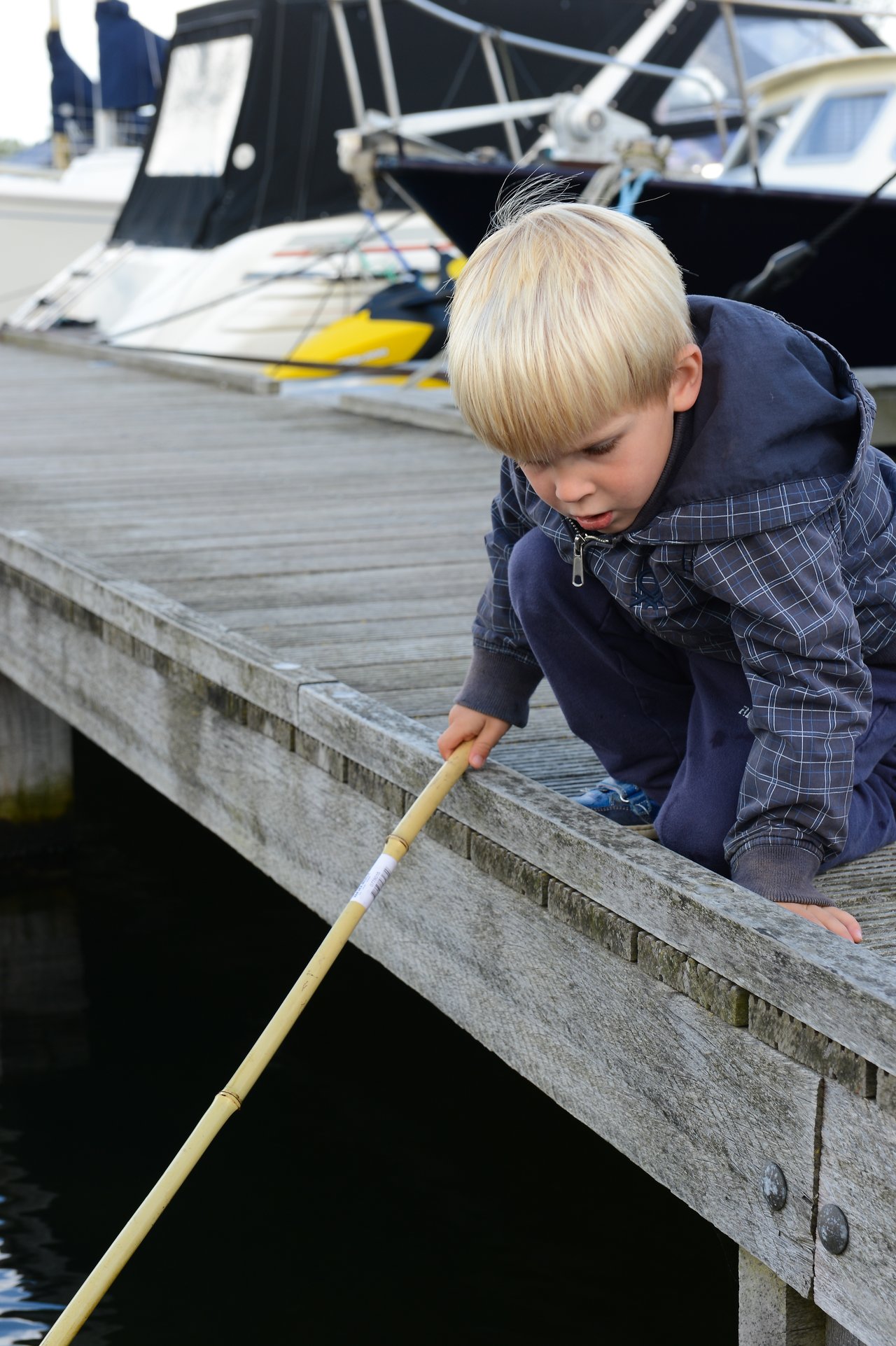 A young child crouches on a wooden dock, holding a long stick over the water, looking down intently.
