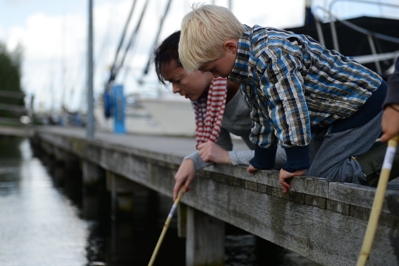 A child and an adult lean over a wooden dock, using sticks to explore the water below.
