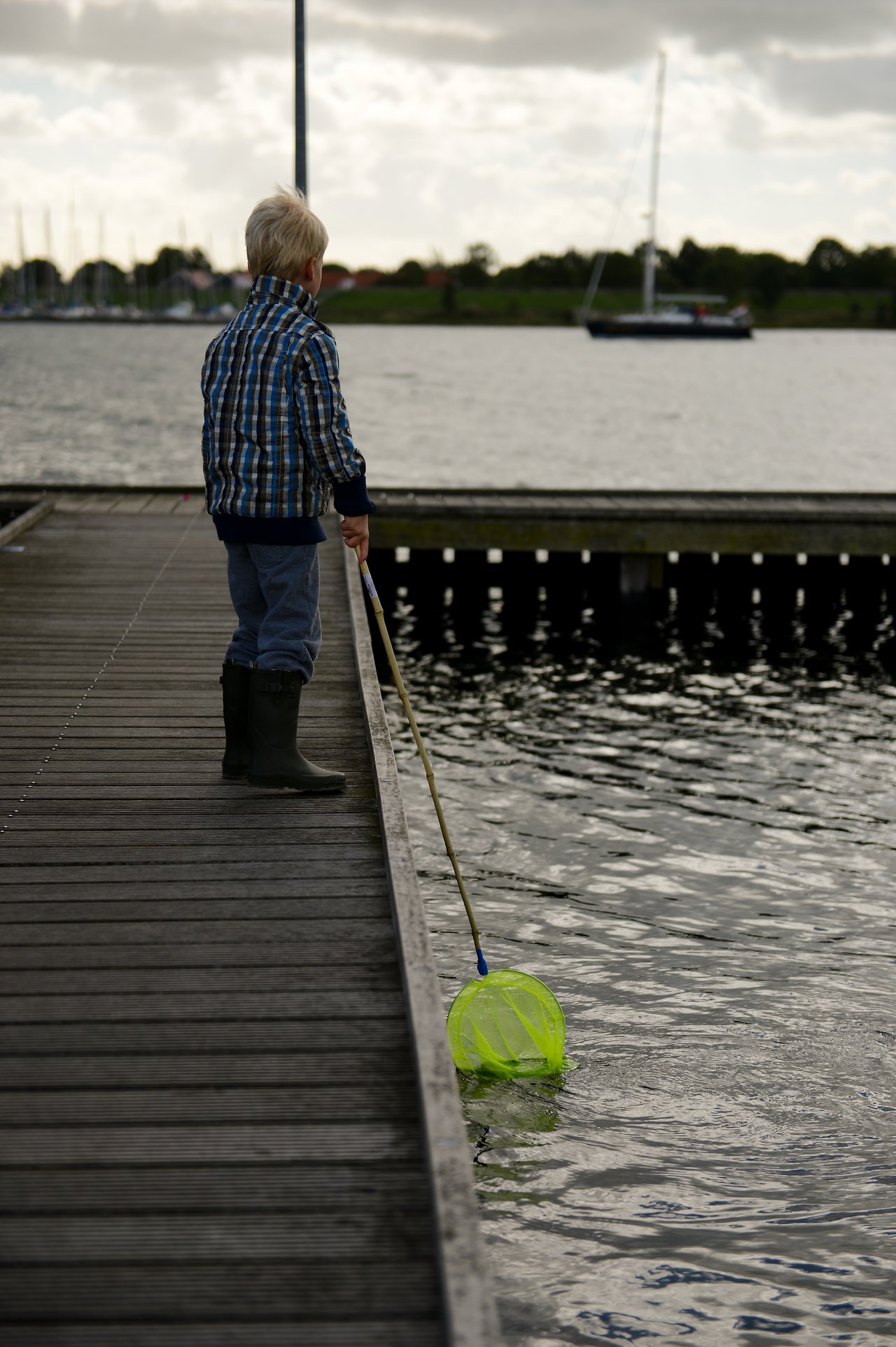 A child stands on a wooden dock, dipping a green fishing net into the water.