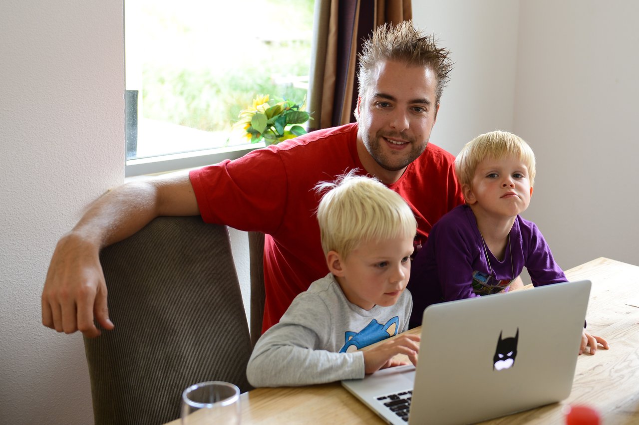 A man and two young children sit at a table, looking at a laptop screen together.