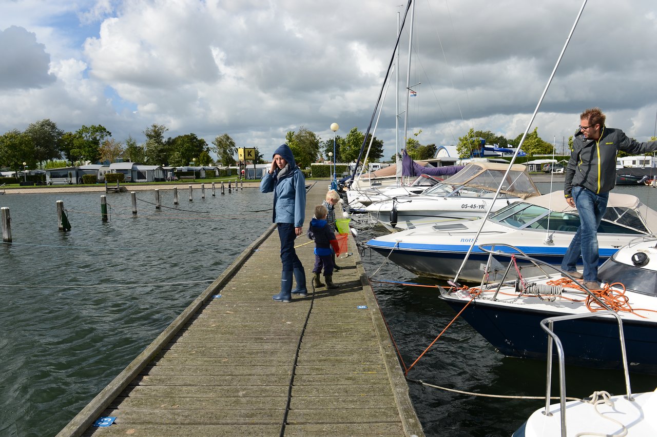 A person in a hooded jacket talks on the phone while children walk on a dock near boats.