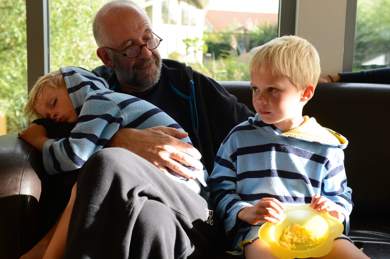 An older man sits on a couch, holding a sleeping child, while another child eats from a yellow bowl.