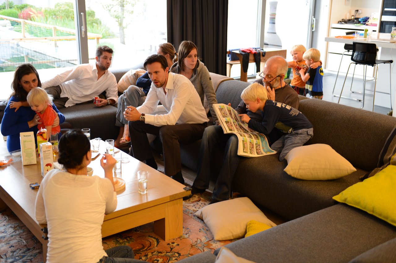 A group of people gathers in a living room, with a child reading a book with an older man.