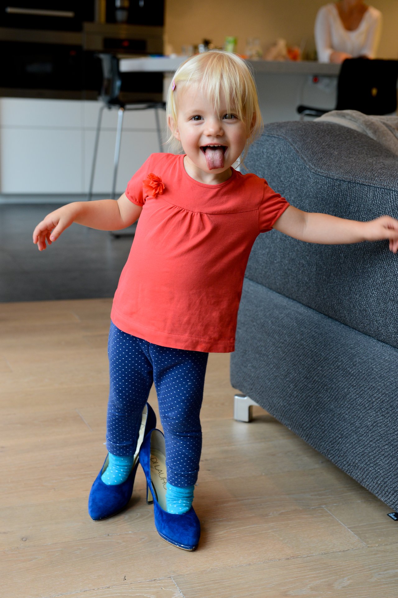 A young child in a red shirt playfully sticks out their tongue while wearing oversized blue high heels indoors.