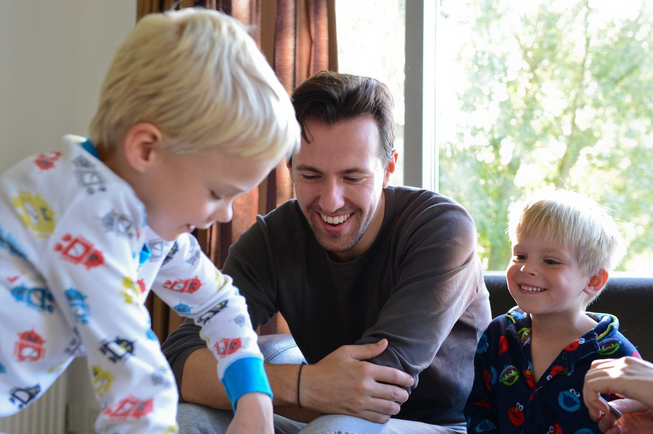 A man and two young children smile and play together indoors near a window with natural light.