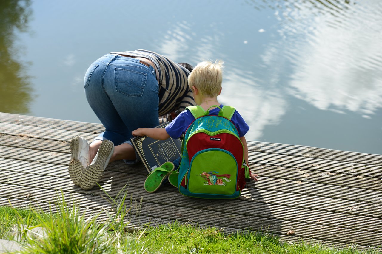 A child with a colorful backpack sits on a wooden dock, watching an adult leaning over the water.