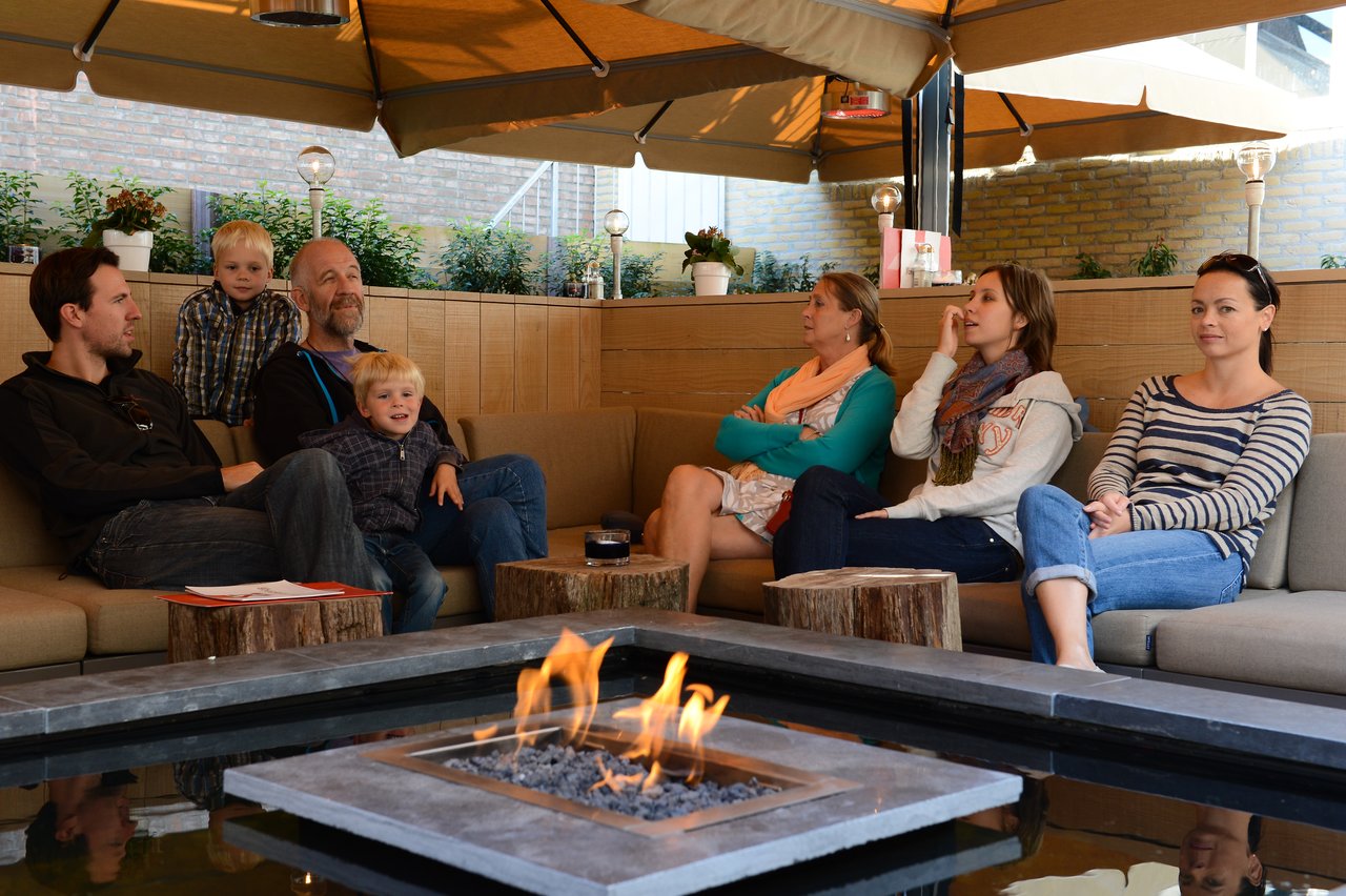 A group of people, including two children, sit around a fire pit under a covered outdoor seating area.