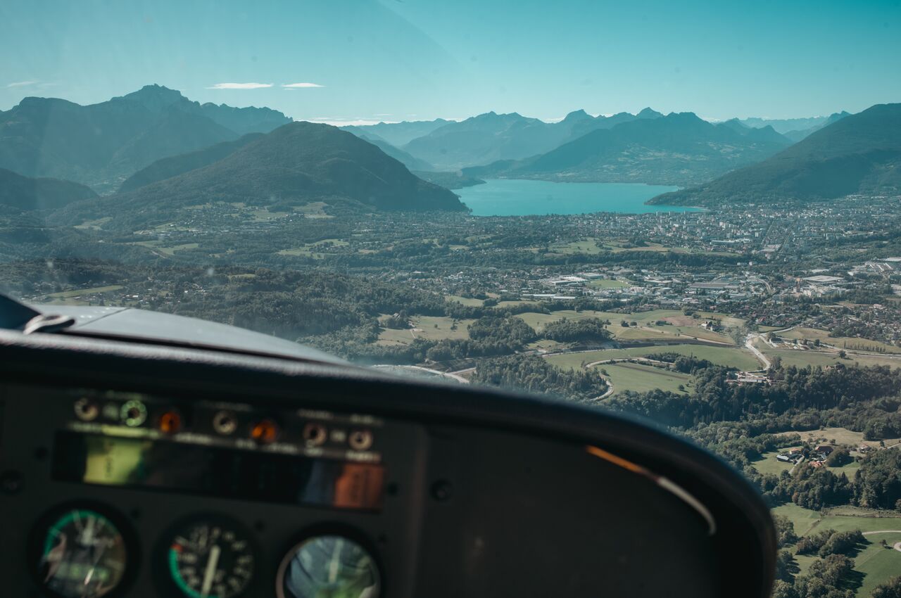 View from a small plane cockpit approaching Annecy, with Lake Annecy and surrounding mountains ahead.