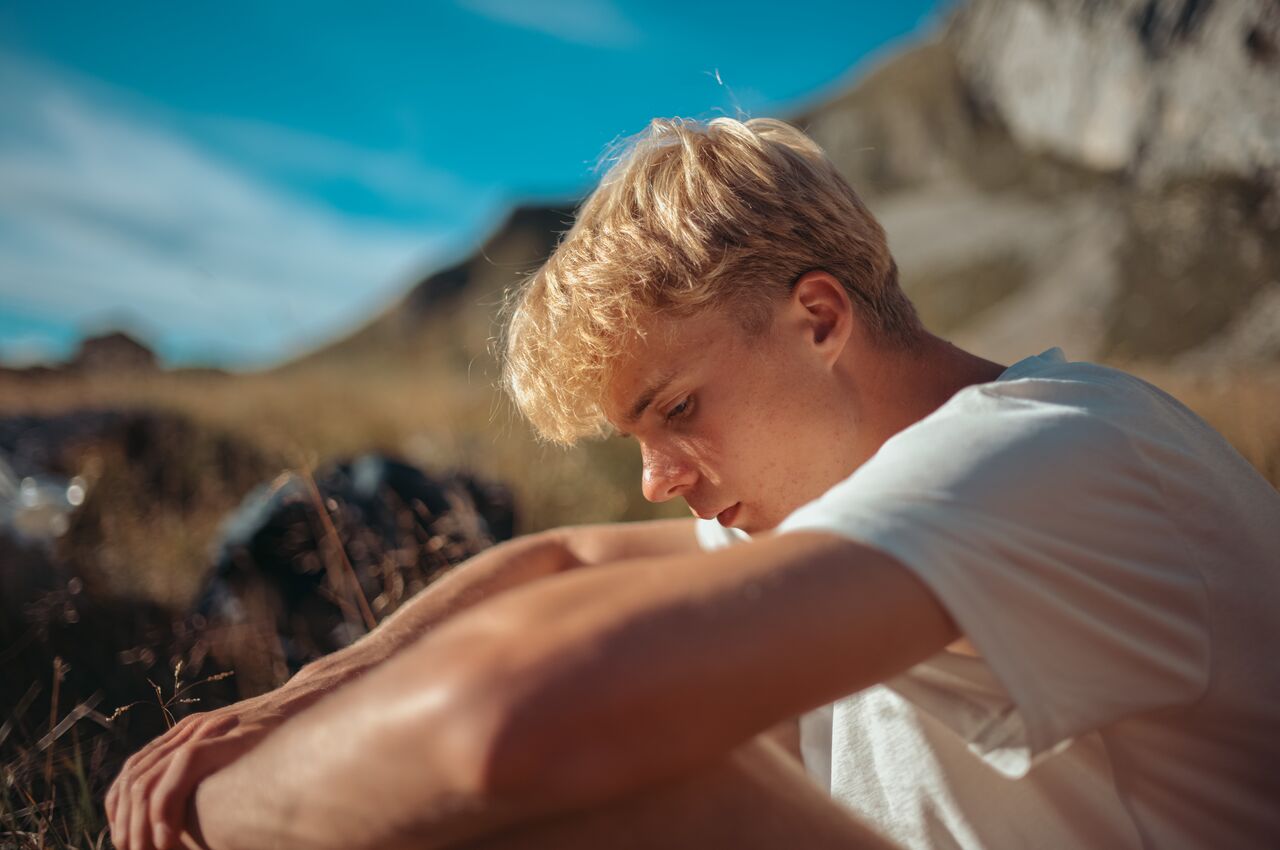 Young man sits on the grass, head down and arms around knees, resting quietly after a long day hiking on a sunny mountain trail.