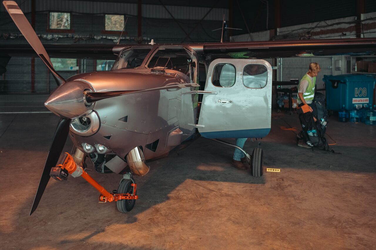 Small Cessna plane in a hangar with its door open, while a person in a safety vest loads backpacks into the cabin, preparing for departure.